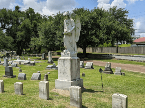 A white angel statue stands on a pedestal in a cemetery, surrounded by gravestones and lush green grass.