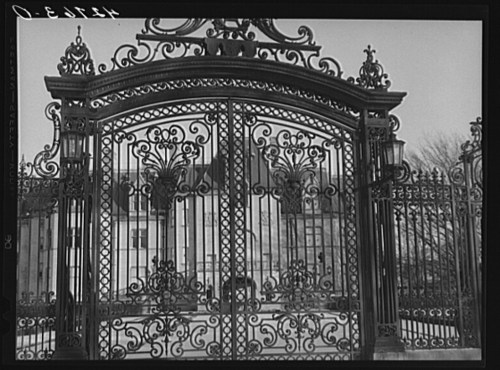 Ornate wrought iron gates with intricate designs, leading to a building in the background.