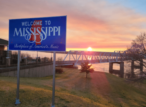 A "Welcome to Mississippi" sign at sunset, with a bridge and river in the background.