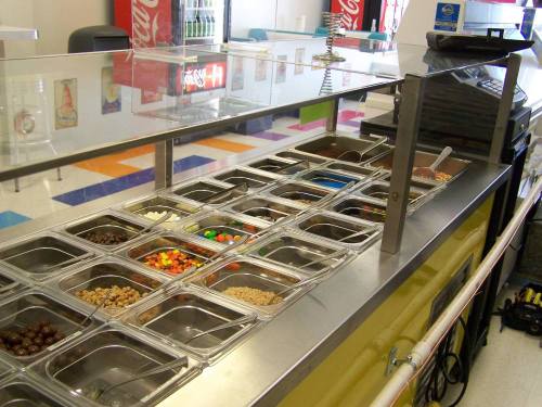 A colorful toppings bar with various containers filled with candies, nuts, and other toppings in a bright setting.