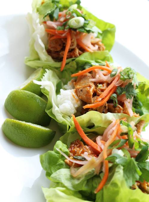 Three lettuce wraps filled with chicken, rice, carrots, and herbs, served with lime wedges on a white plate.