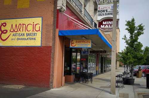 Exterior of Essencia Artisan Bakery with a blue awning, outdoor seating, and nearby shops in a city setting.