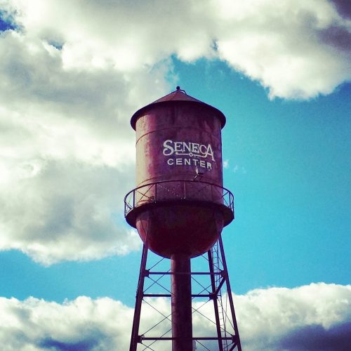Rusty water tower labeled "Seneca Center" against a backdrop of blue sky and fluffy clouds.