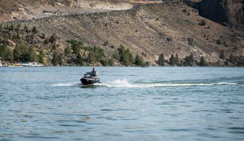 A person rides a jet ski on a calm lake, surrounded by hills and trees in the background.