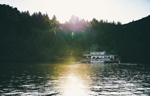 A houseboat floats on a calm lake, surrounded by trees and mountains, with sunlight reflecting on the water.