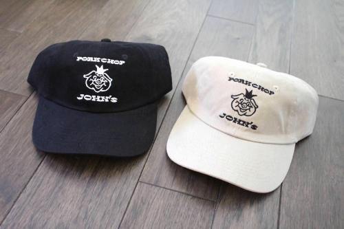 Two baseball caps, one black and one beige, both featuring the text "PORK CHOP JOHN'S" and a cartoon pig logo.