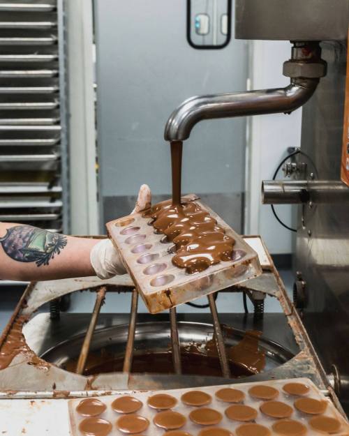 A hand pours melted chocolate into a mold in a kitchen, with trays of chocolate in the background.