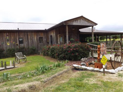 A rustic wooden building with a porch, surrounded by greenery and flowers, featuring a wagon wheel and a caution sign.