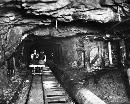 Two miners sit on a cart in a dimly lit underground tunnel, surrounded by rock and wooden supports.