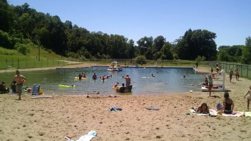 A sunny beach scene with people swimming and relaxing by a pond, surrounded by green trees and sandy areas.