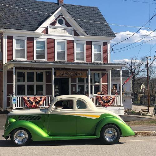 A vintage green and yellow car parked in front of a red and white general store with bunting decorations.