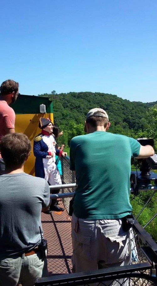 A film crew captures a scene outdoors with a person in costume against a scenic green backdrop.