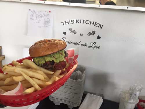 A hand holds a burger topped with guacamole and fries in a red basket, with a sign reading "This kitchen is seasoned with love."