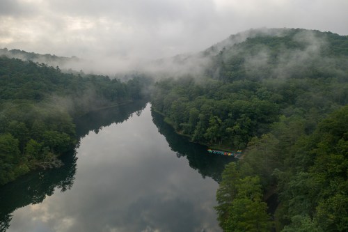 A serene river winds through lush green hills, shrouded in mist, with colorful kayaks visible on the water.
