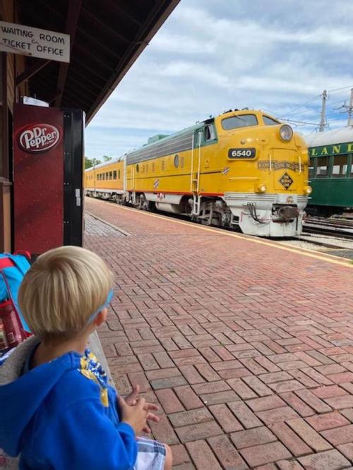 A child sits on a bench at a train station, watching a yellow vintage train approach. A Dr Pepper vending machine is nearby.