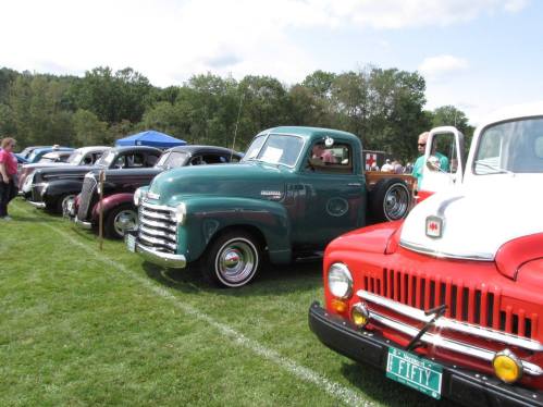 A row of classic cars and trucks parked on grass, showcasing vintage designs and vibrant colors under a blue sky.