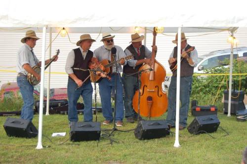 A bluegrass band performs under a tent, featuring five musicians with instruments like banjo, fiddle, and double bass.