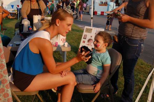 A young girl sits in a chair while a woman applies face paint to her cheek at an outdoor event.