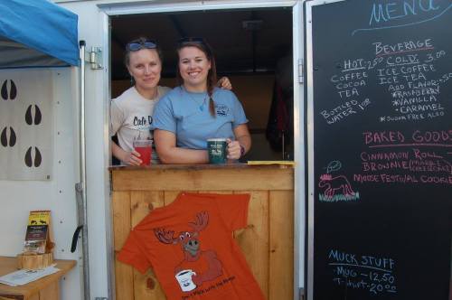 Two women smile from a food truck window, holding mugs, with a menu and a moose-themed shirt displayed nearby.