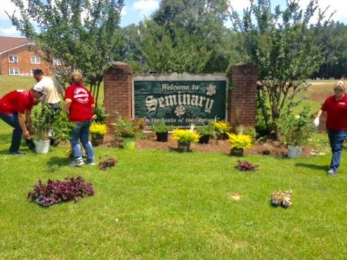 People planting flowers and shrubs around a "Welcome to Seminary" sign in a green, landscaped area.
