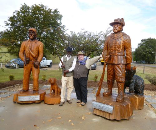 A man poses between two wooden statues of historical figures, with a dog statue at their feet in a park setting.