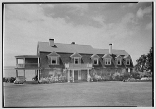 A large, two-story house with a gabled roof, surrounded by greenery and a clear sky in the background.