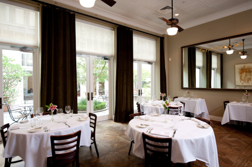 Elegant restaurant interior with white tablecloths, wooden chairs, large windows, and greenery visible outside.