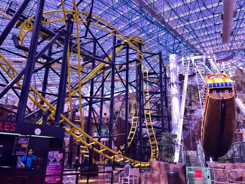 Indoor amusement park featuring a yellow and black roller coaster and a pirate ship ride under a glass ceiling.