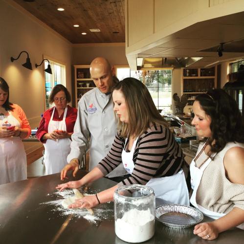 A group of people in an indoor kitchen, learning to bake with a chef, flour scattered on the countertop.