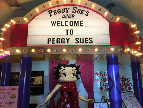 Sign for Peggy Sue's Diner with a welcoming message and a Betty Boop statue in front. Brightly lit and colorful decor.