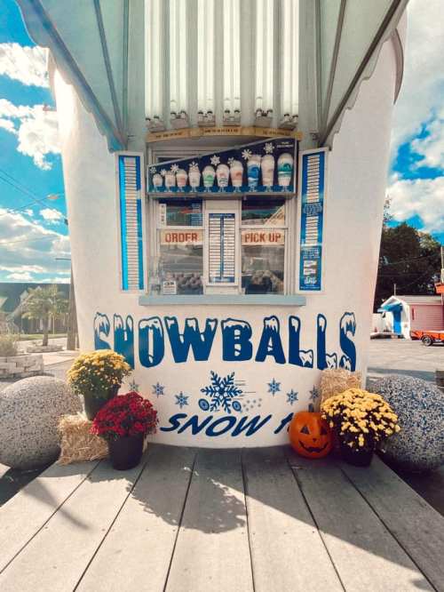 A colorful snowball stand with a menu, flowers, and a pumpkin outside, set against a bright blue sky.