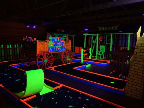 A vibrant glow-in-the-dark mini golf course themed like the Wild West, featuring a wagon, sheriff's office, and colorful pathways.