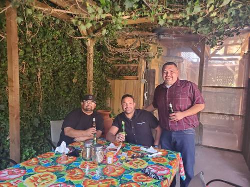 Three men sit at a colorful table under a leafy canopy, each holding a drink and smiling.