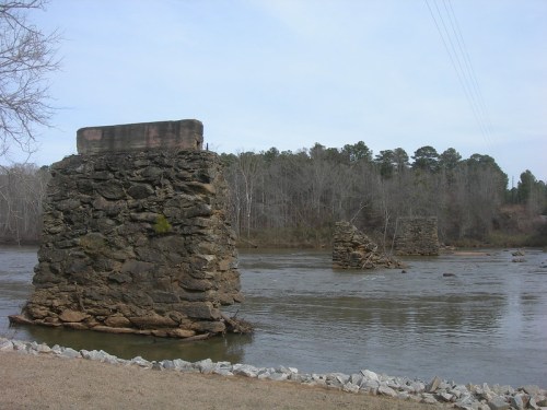 Ruins of stone bridge pillars in a river, surrounded by trees and a grassy bank under a cloudy sky.