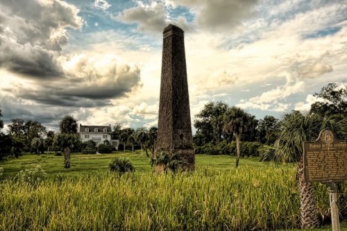 A tall stone monument stands in a grassy field, with a historic house and dramatic clouds in the background.