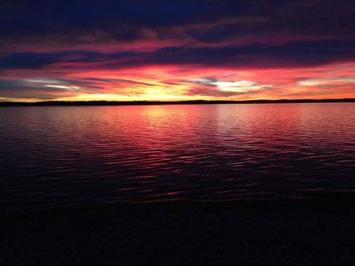 A vibrant sunset over a calm lake, with colorful reflections on the water and a darkening sky.