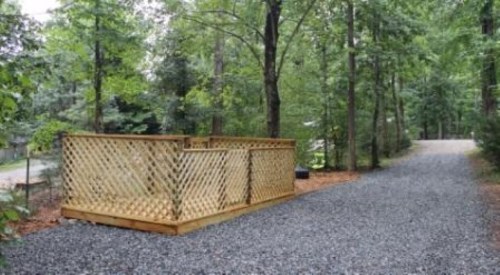 A wooden deck with lattice walls sits beside a gravel path in a wooded area. Trees surround the space.