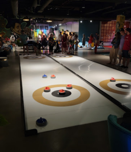A lively indoor curling area with players and spectators, featuring two lanes with target circles and colorful stones.