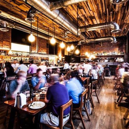 A bustling restaurant with wooden decor, dim lighting, and patrons enjoying meals at tables.
