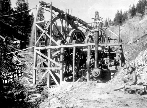 Historic black-and-white photo of the Taylor Mill and Mine, featuring large machinery and a worker seated nearby, 1893.