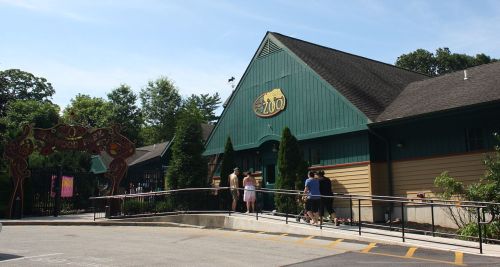 A green and brown zoo building with visitors lined up outside on a sunny day.