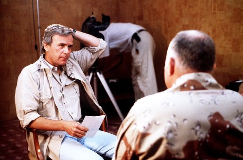 A man in a beige shirt sits in an interview setting, facing another man in a patterned shirt, with a camera in the background.
