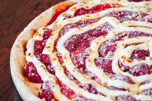 A close-up of a dessert pizza topped with red fruit, cream, and a dusting of powdered sugar on a wooden surface.