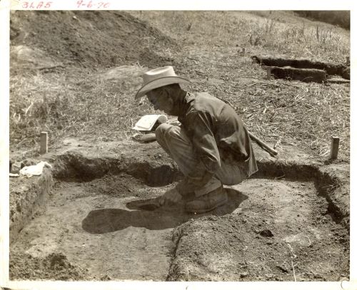 A man in a hat crouches over a small excavation site, examining the ground with a notebook in hand.
