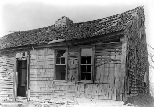 A dilapidated, weathered house with a slanted roof and broken windows, surrounded by a barren landscape.