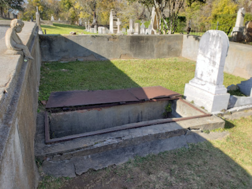 A grave site with a metal cover and a headstone, surrounded by grass and other gravestones in a cemetery.