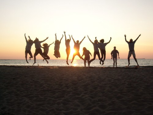 A group of people jumping joyfully on a beach at sunset, silhouetted against the glowing horizon.