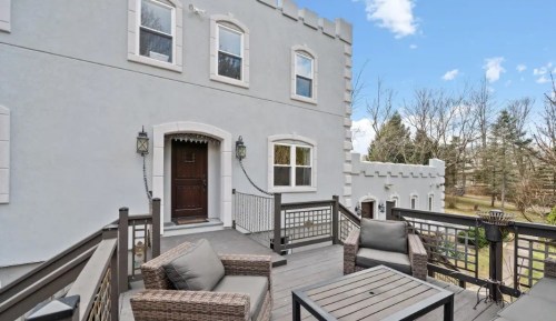A cozy outdoor deck with wicker furniture, overlooking a landscaped area and a light-colored building with castle-like features.