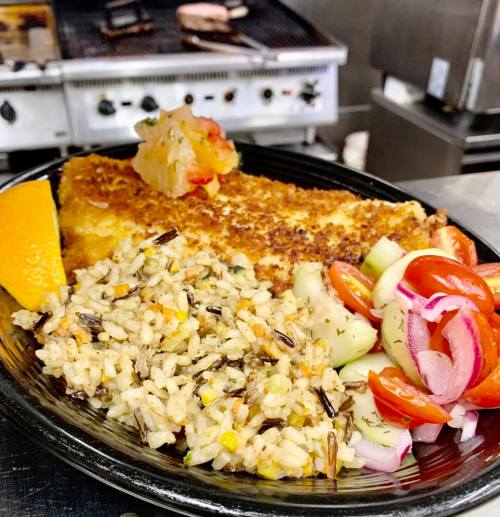 A plate featuring crispy fish, wild rice, and a fresh salad with tomatoes and cucumbers, garnished with an orange slice.