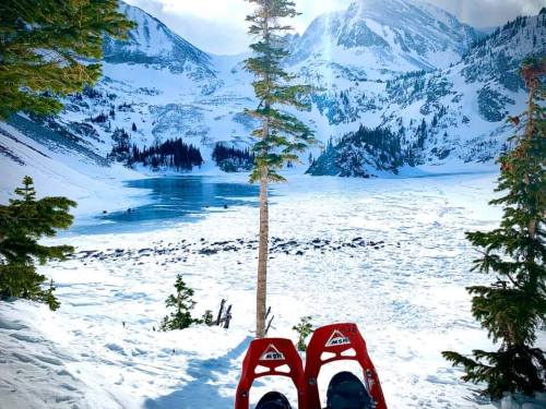 Snow-covered landscape with mountains, a frozen lake, and snowshoes in the foreground. Bright, sunny atmosphere.
